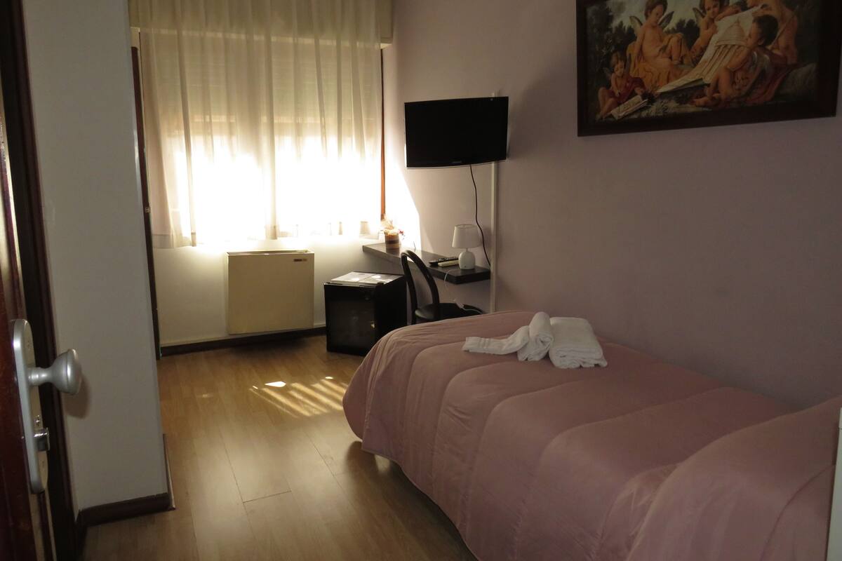 A hotel room with a bed, a desk, a chair, a television, a painting, and a window with curtains.