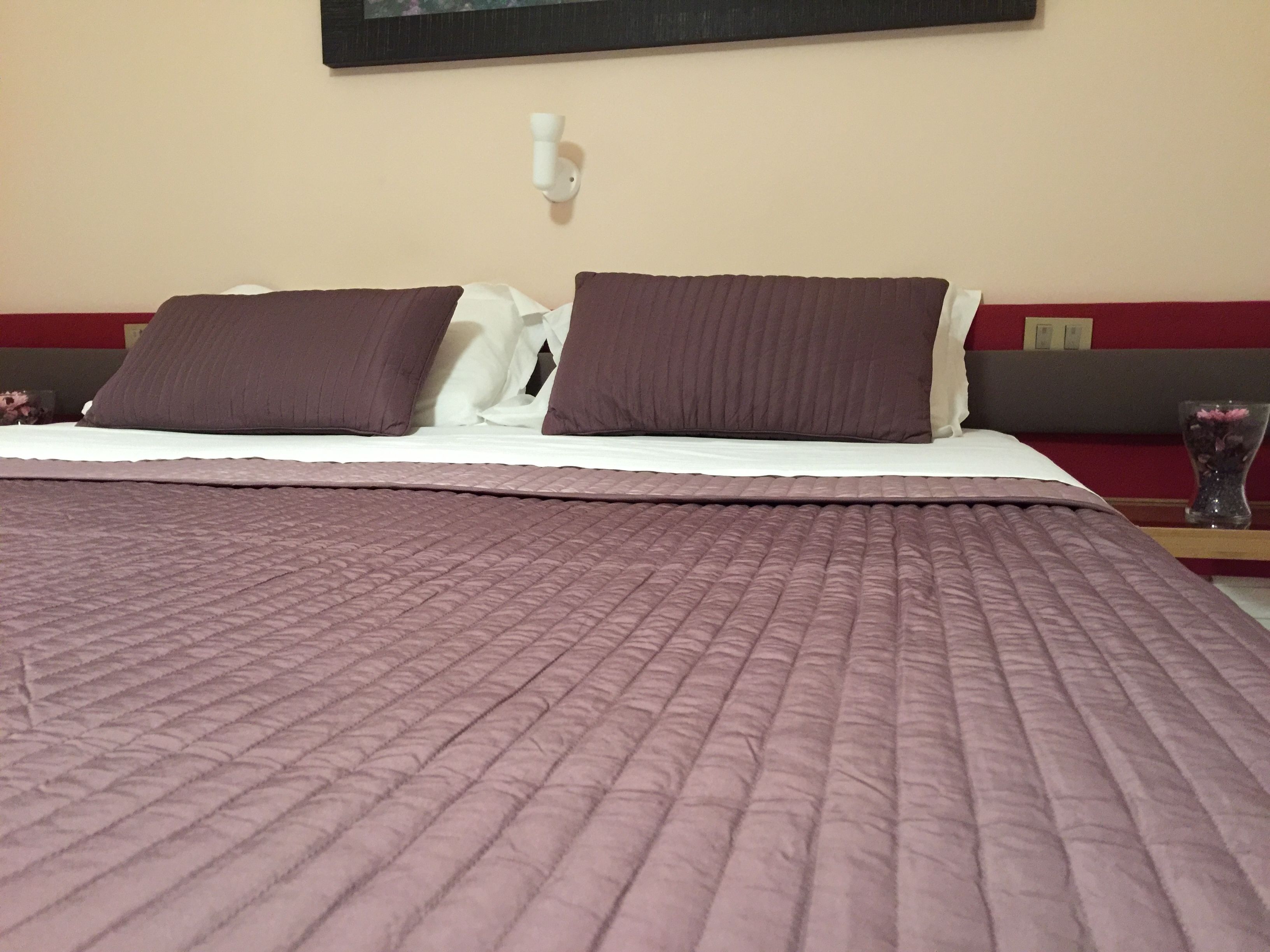 A neatly made bed with a purple quilt, two dark pillows, and a white pillow.