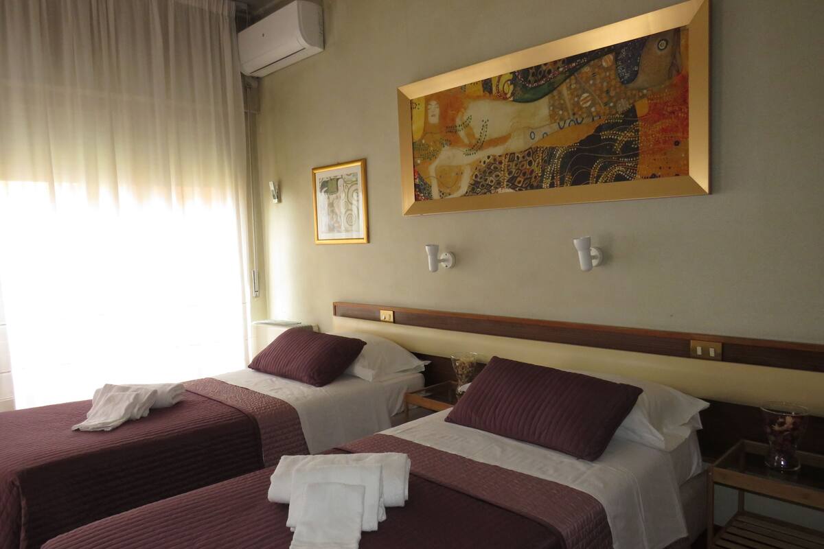 A hotel room with two beds, each with a white and maroon bedspread, and a painting above the beds.