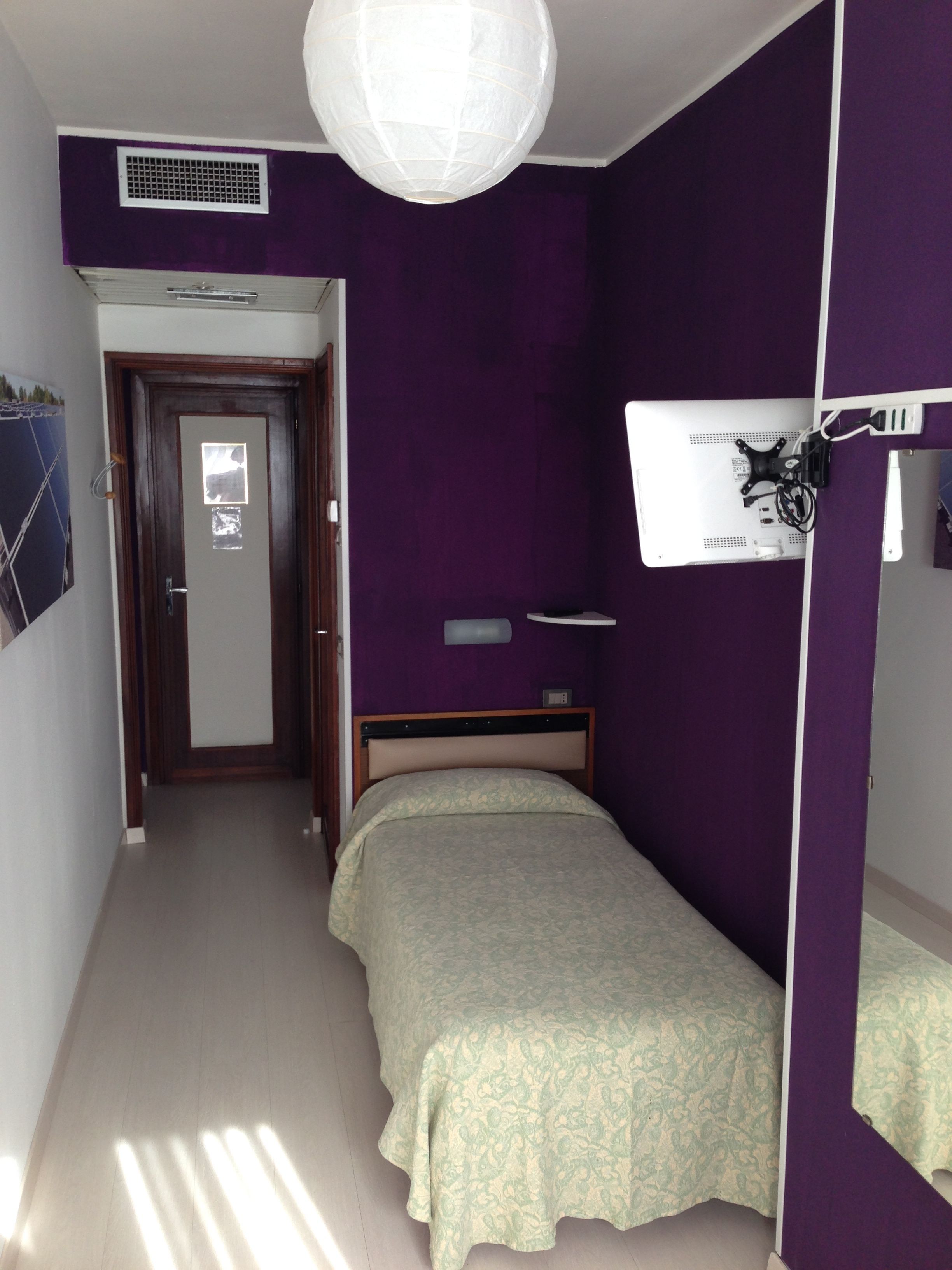 A bedroom with purple walls, a bed with a patterned bedspread, a white ceiling light, and a door leading to another room.
