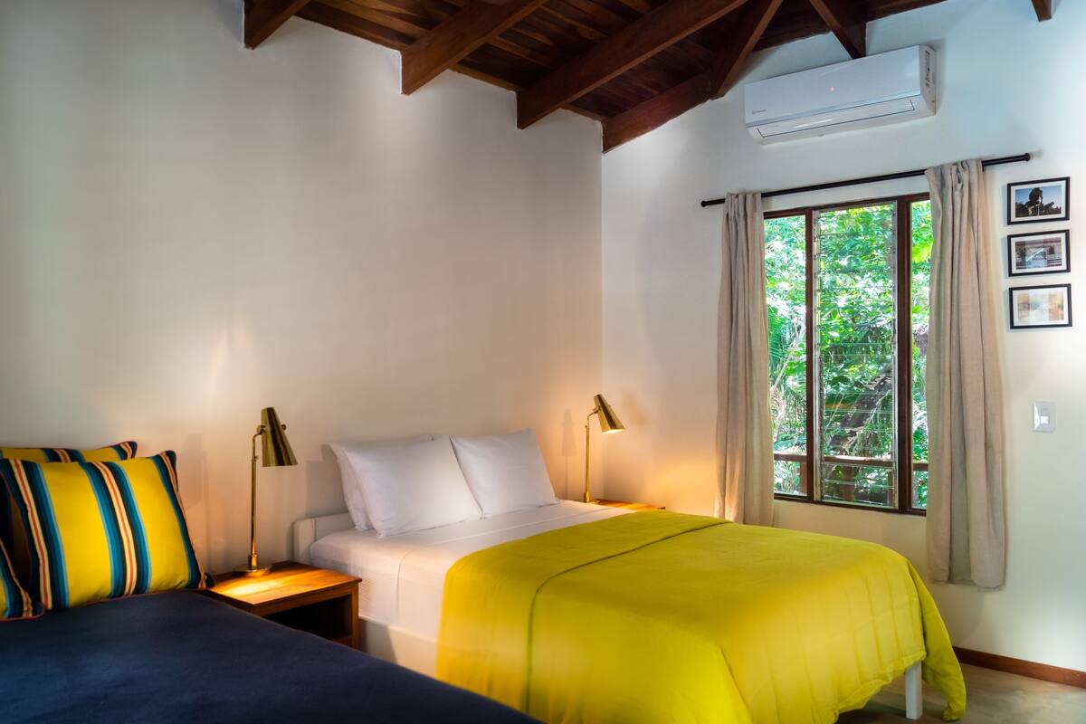 A room with two beds, a window with curtains, an air conditioner, and a wall-mounted picture.