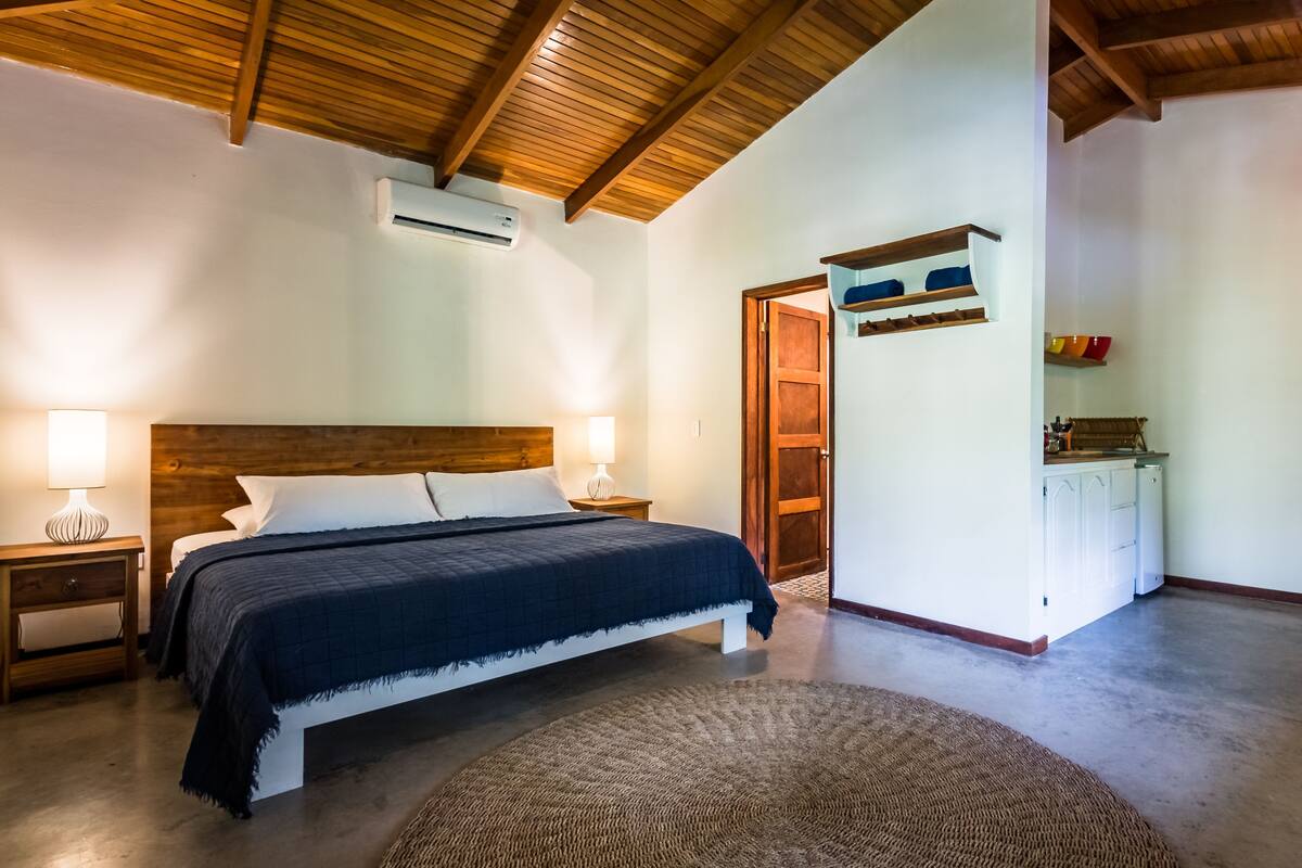 A bedroom with a wooden ceiling, a bed with a dark blanket, bedside lamps, a white wardrobe, and a small kitchen area.