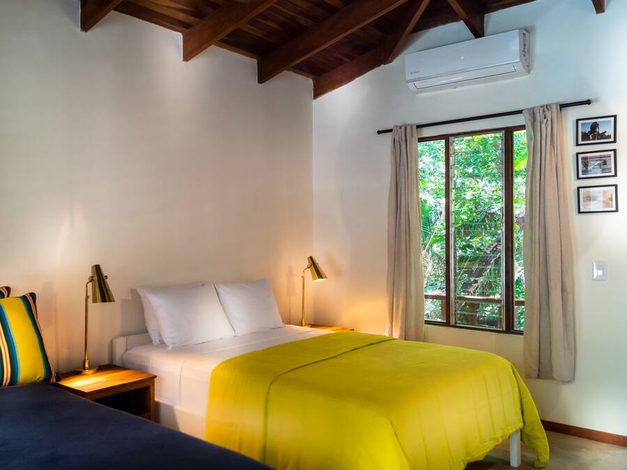 Quarto com duas camas, janela com cortinas e aparelho de ar condicionado.