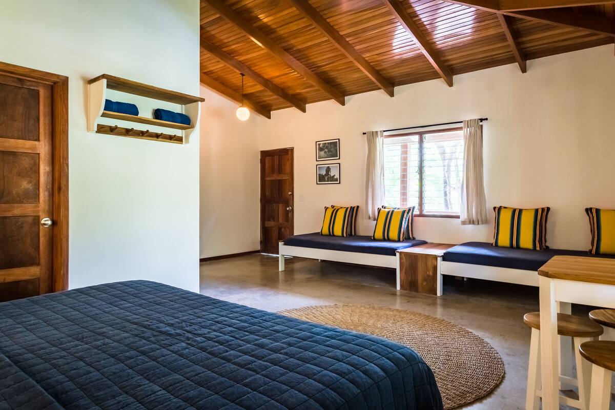 A room with a bed, two seating areas with cushions, a wooden ceiling, a door, and a window with curtains.