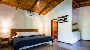Un dormitorio con techo de madera, cama con cubrecama azul oscuro, dos lámparas de noche, un armario blanco y un pequeño estante con adornos.