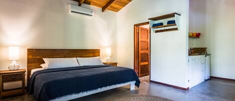 A bedroom with a wooden ceiling, a bed with a dark blue bedspread, two bedside lamps, a white wardrobe, and a small shelf with decorative items.