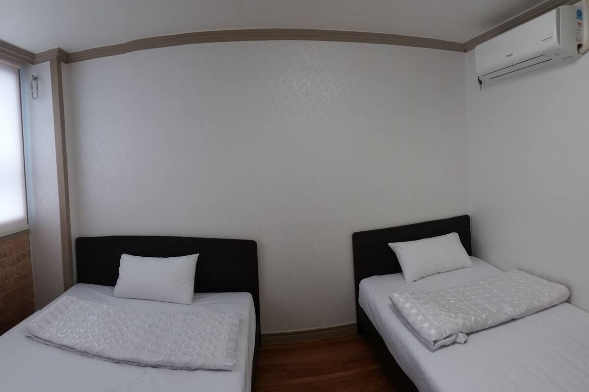 A room with two single beds, white bedding, a brick wall, and an air conditioning unit.