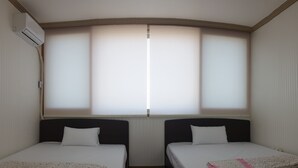 Habitación con dos camas, cada una con colchón blanco y manta estampada con motivos florales. Se observa una ventana con persianas, y un equipo de aire acondicionado instalado en la pared.