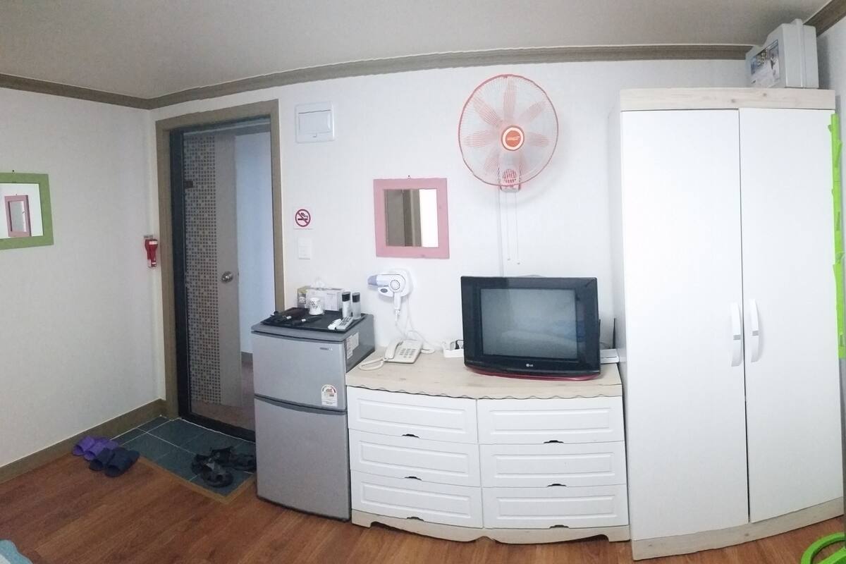 A small room with a refrigerator, a microwave, a small cabinet with drawers, a television, a fan, and a door with a mirror.
