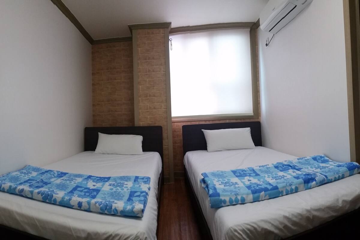 A room with two beds, each with a blue floral bedspread and white pillows. There is a brick wall and a window with a blind.
