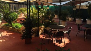 Un espacio al aire libre con patio, con una mesa redonda y sillas, rodeado de plantas en macetas y una valla verde.