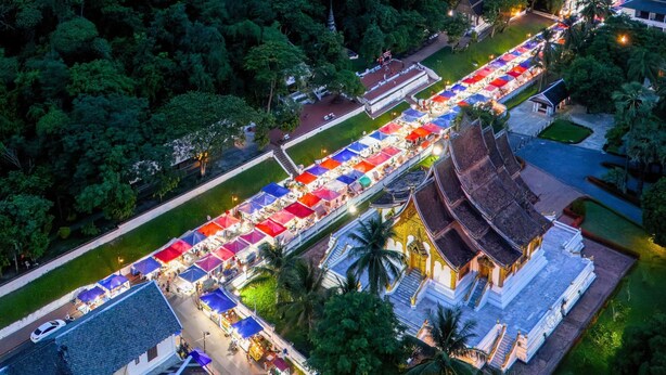 An aerial view of a street lined with colorful market stalls, a traditional temple, and surrounding greenery.