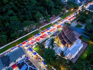 An aerial view of a street lined with colorful market stalls, a traditional temple, and surrounding greenery. - 3 Nagas Hotel Luang Prabang - MGallery Collection (Luang Prabang)