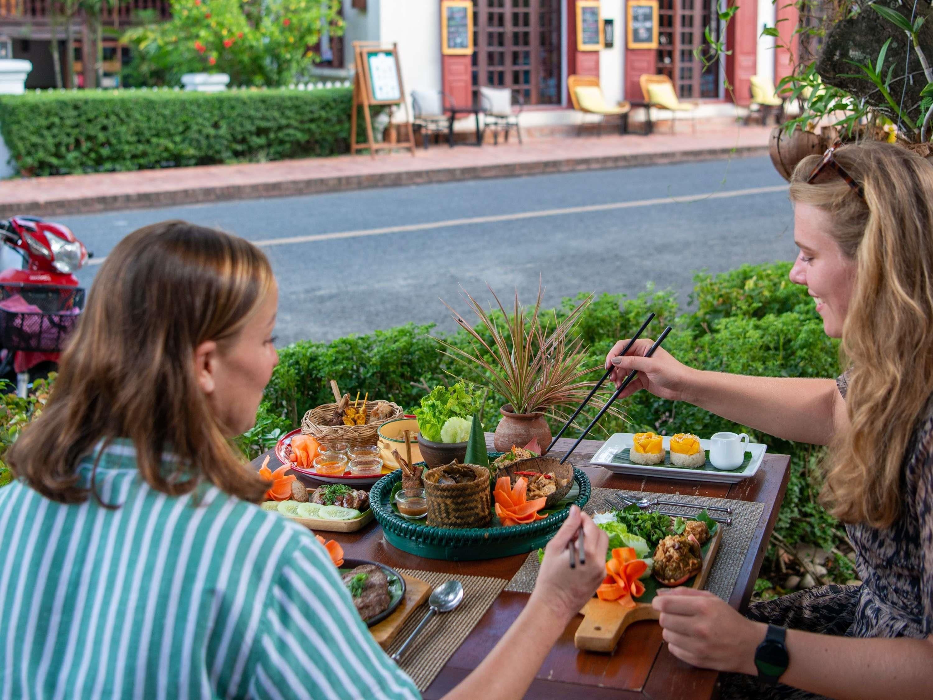 Two people dining outdoors with a variety of dishes and a table set with chopsticks and a teapot.