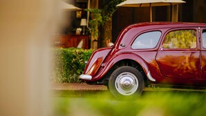 A vintage red car parked in a lush garden area with a wooden building and large umbrellas in the background. - 3 Nagas Hotel Luang Prabang - MGallery Collection (Luang Prabang)