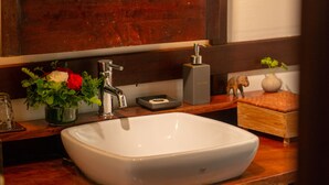 A bathroom with a wooden vanity, a white sink, a mirror, and a decorative vase with flowers. - 3 Nagas Hotel Luang Prabang - MGallery Collection (Luang Prabang)