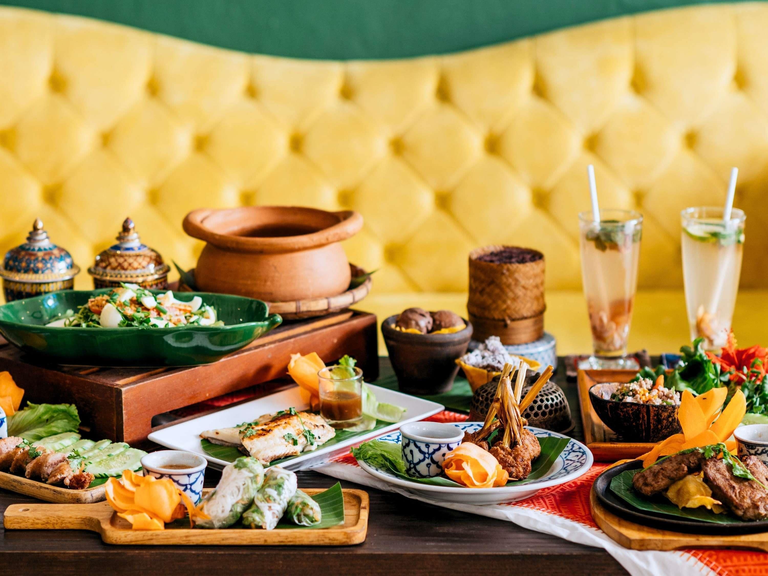 A table set with various dishes, including a salad, grilled meat, and desserts, in a room with a yellow upholstered headboard.