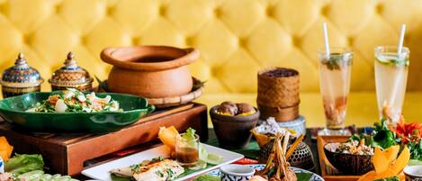 A table set with various dishes, including a salad, grilled meat, and desserts, in a room with a yellow upholstered headboard.