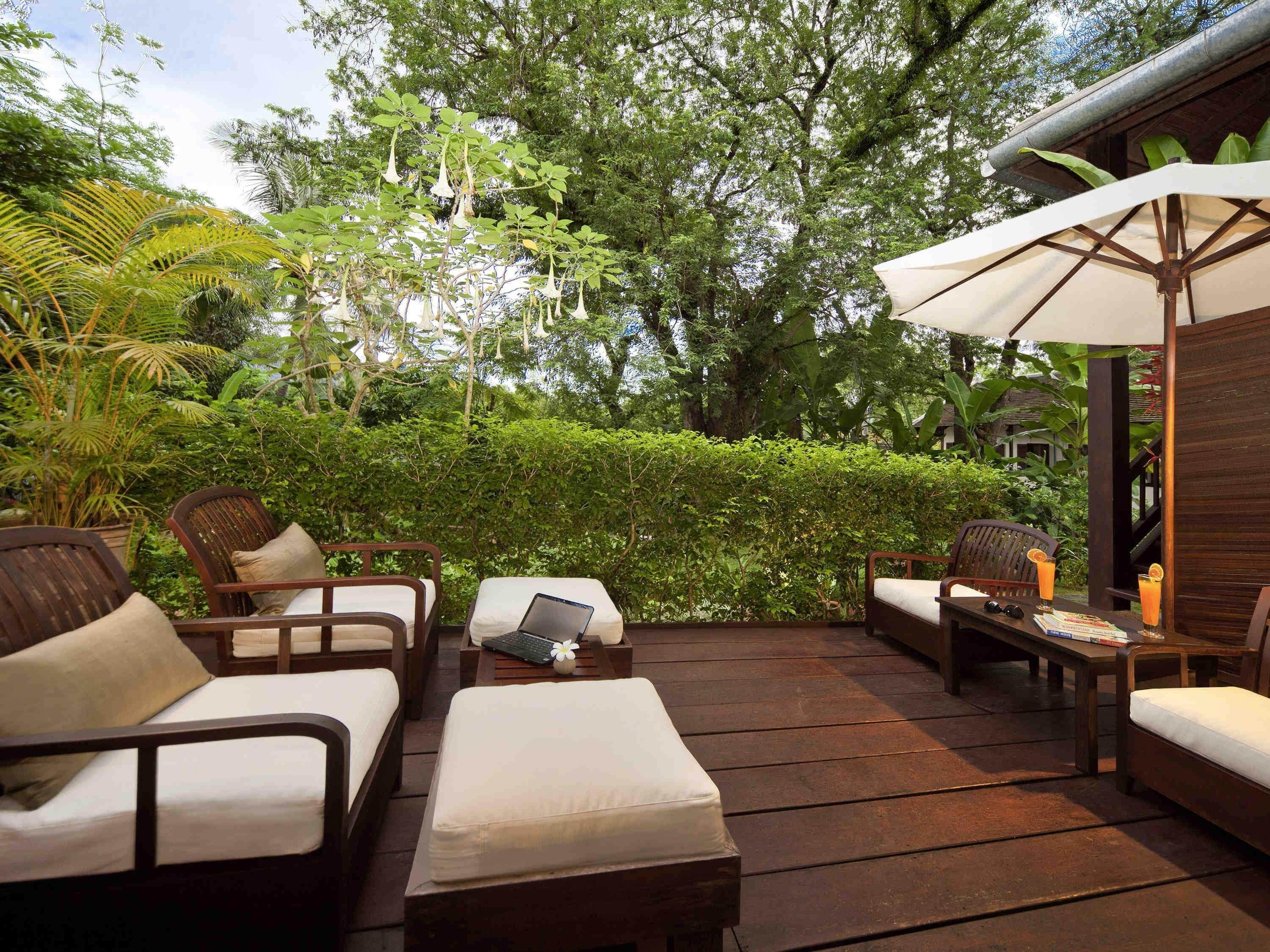 A wooden deck with outdoor furniture, including sofas and a table, surrounded by lush greenery and trees.