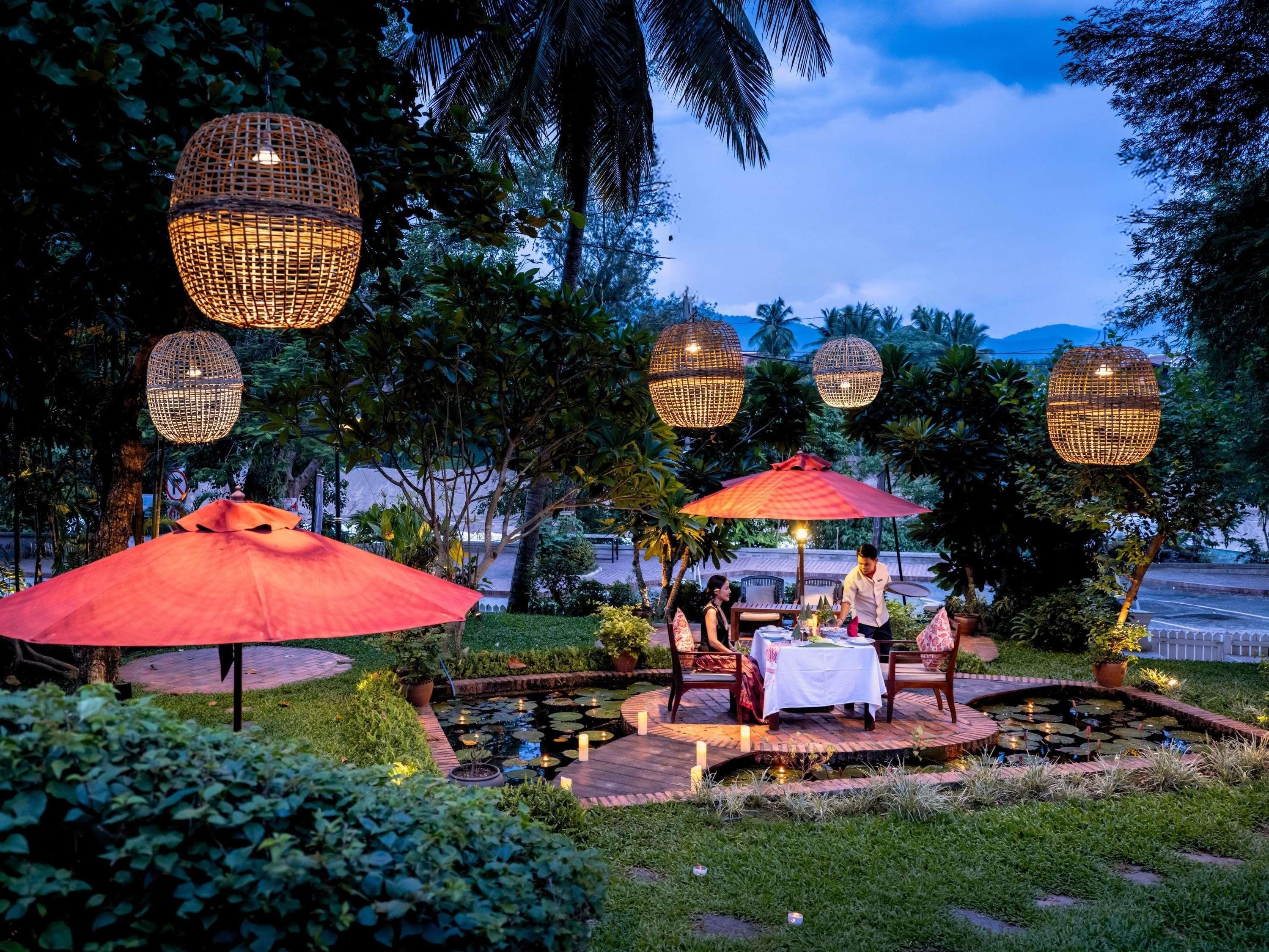 Un área de comedor al aire libre con mesas y sillas, rodeada de un jardín con un estanque e iluminación decorativa.