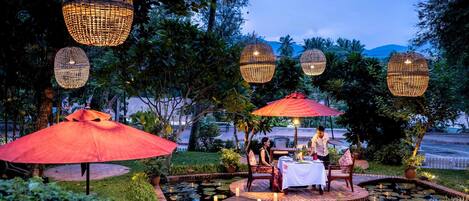Un área de comedor al aire libre con mesas y sillas, rodeada de un jardín con un estanque e iluminación decorativa.