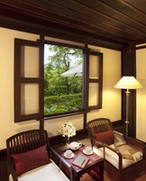 A room with a wooden ceiling, a window with open shutters, a table with a teapot, cups, and a notebook, two chairs with cushions, and a lamp.