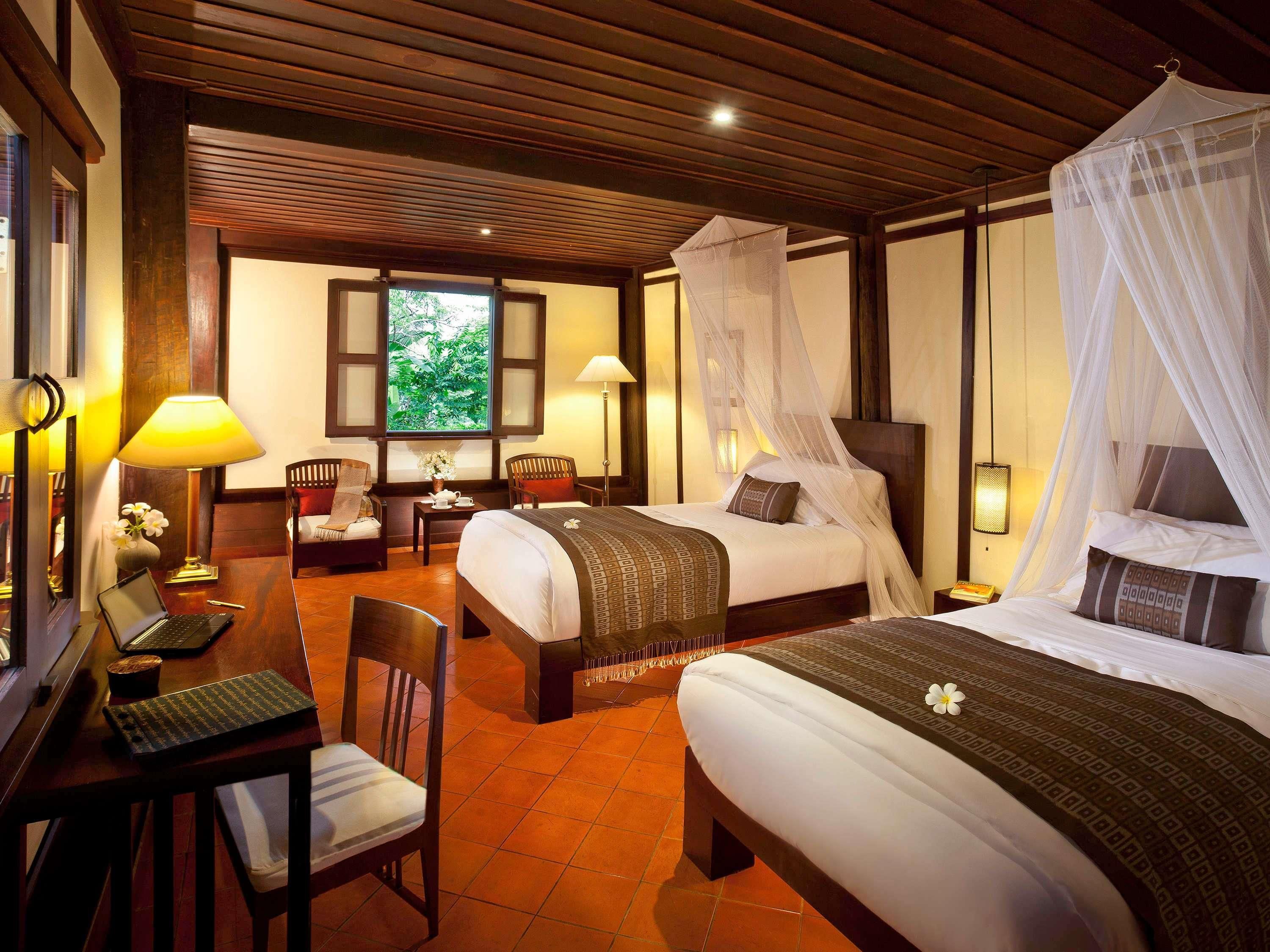 A hotel room with a bed, a desk with a laptop, a chair, a window with a view of greenery, and a canopy bed.