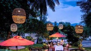 An outdoor dining area with tables and chairs, surrounded by a garden with a pond and decorative lighting.
