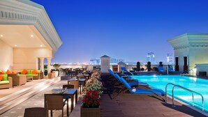 Un área de piscina en la azotea con sillones de descanso, un comedor y vistas del horizonte urbano al atardecer.