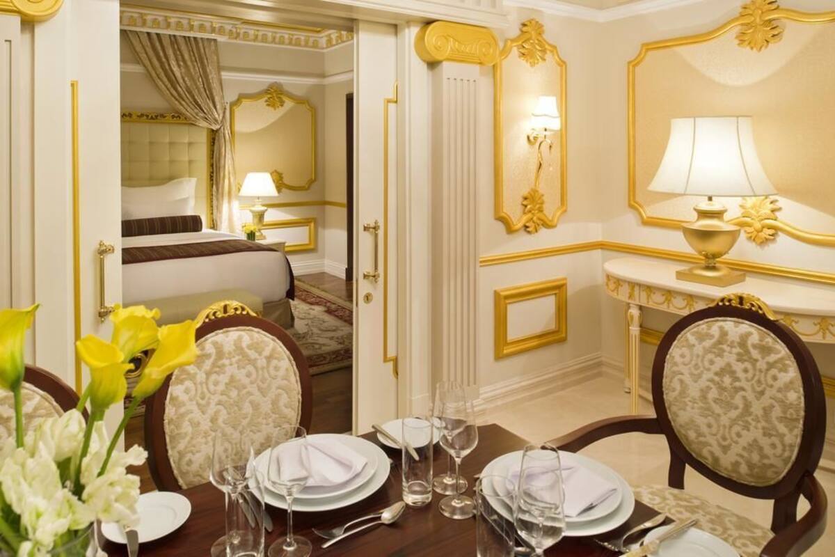 A luxurious hotel room with a neatly made bed, a dining table set for four, and a sitting area with a sofa and a side table.