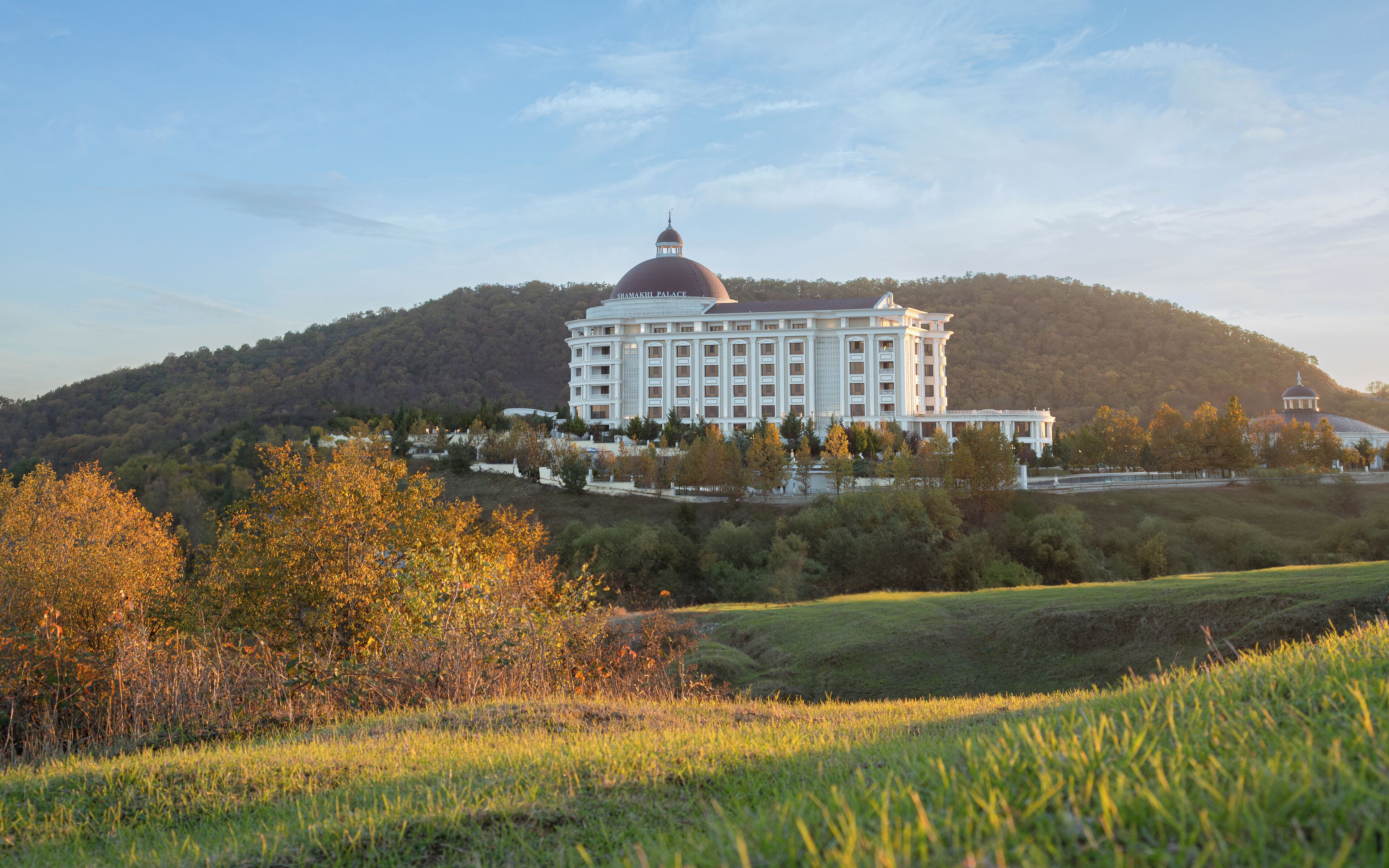 A large, white building with a dome and multiple floors, surrounded by trees and hills.