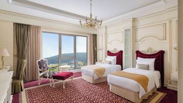A hotel room with two beds, a chandelier, a large window with a view, and a patterned carpet.