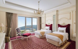A hotel room with two beds, a chandelier, a large window with a view, and a patterned carpet.