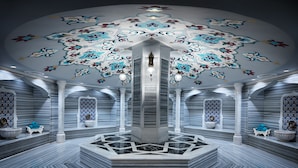 Un baño lujoso con una fuente central, pisos de mármol y elaborada decoración en el techo.