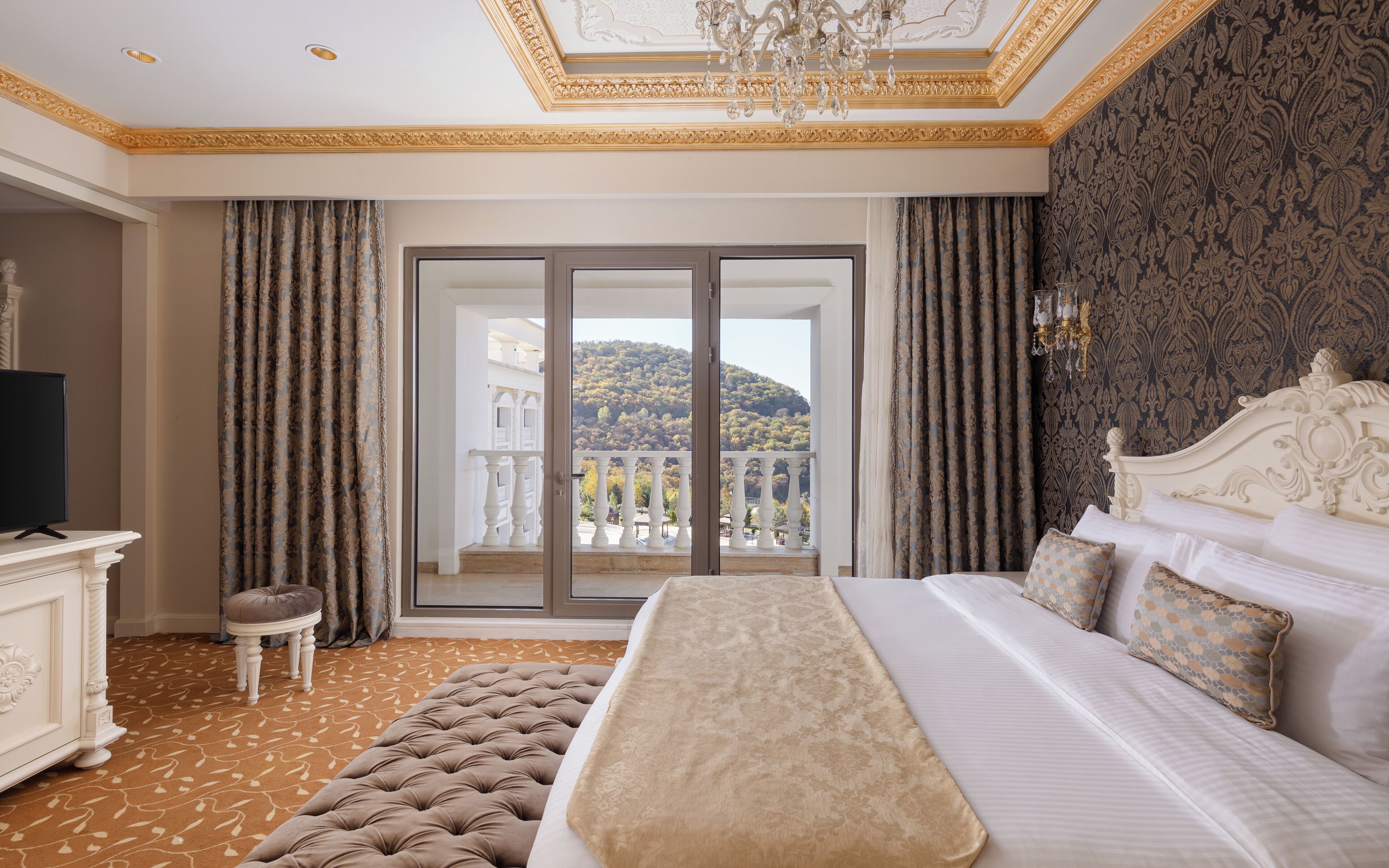A hotel room with a large bed, a television, a small table, and a view of a balcony with a mountain backdrop.