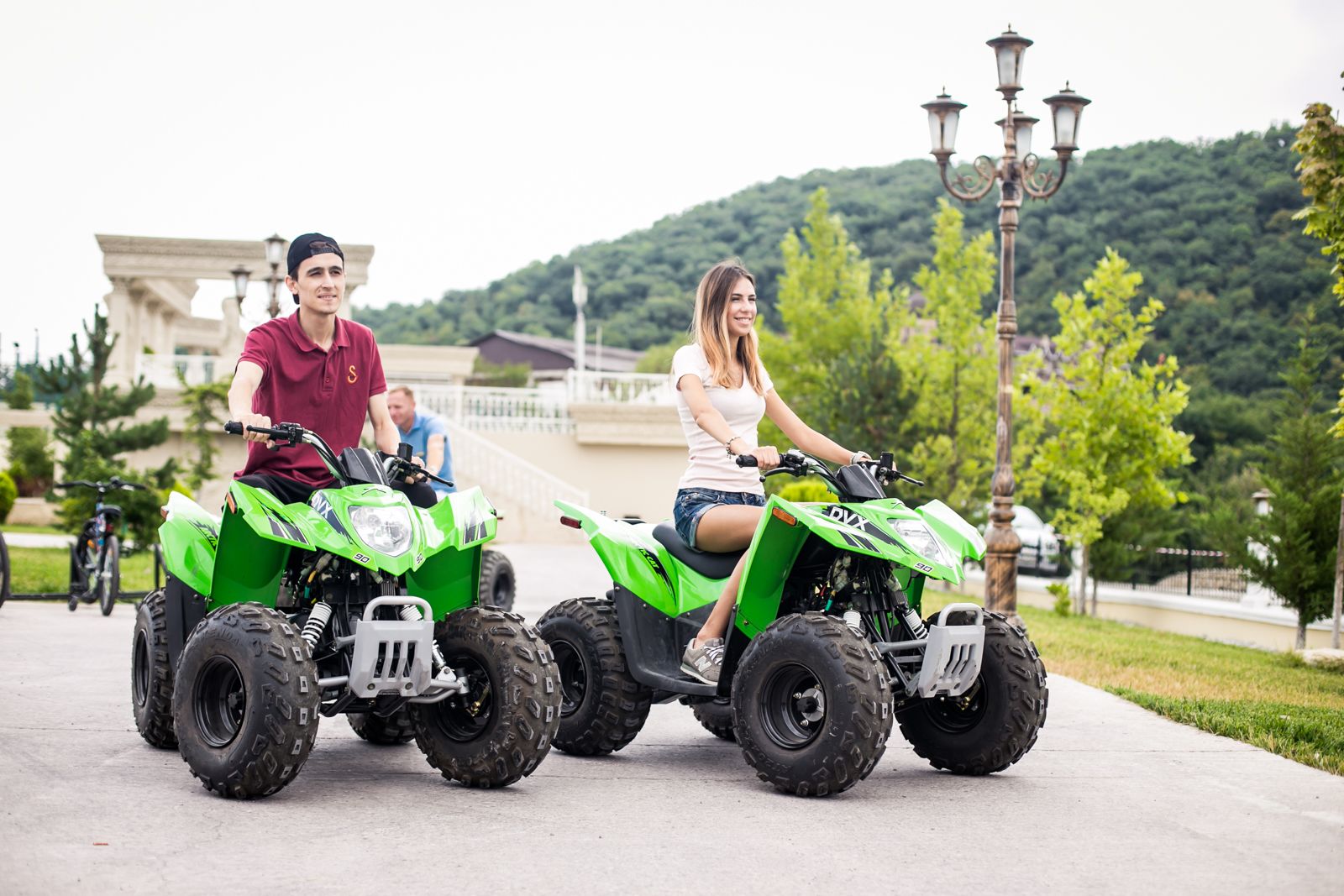 Two people riding green all-terrain vehicles on a paved path with a building and trees in the background.