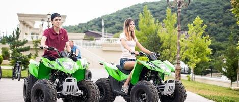 Two people riding green all-terrain vehicles on a paved path with a building and trees in the background.