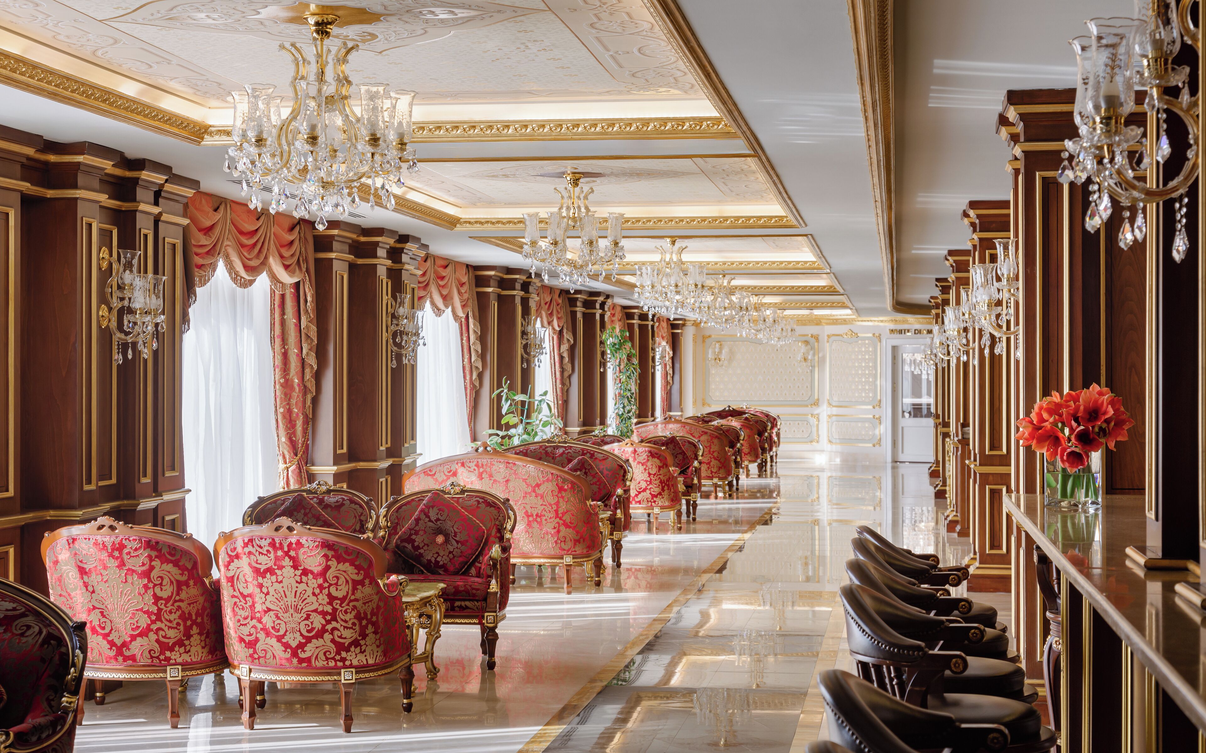 Un lobby de hotel lujoso con candelabros ornamentados, sillones tapizados en rojo y un piso de mármol pulido.
