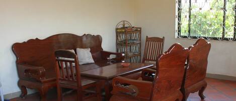 A dining area with a wooden table and chairs, a small shelf with books, and a window with a metal grill.