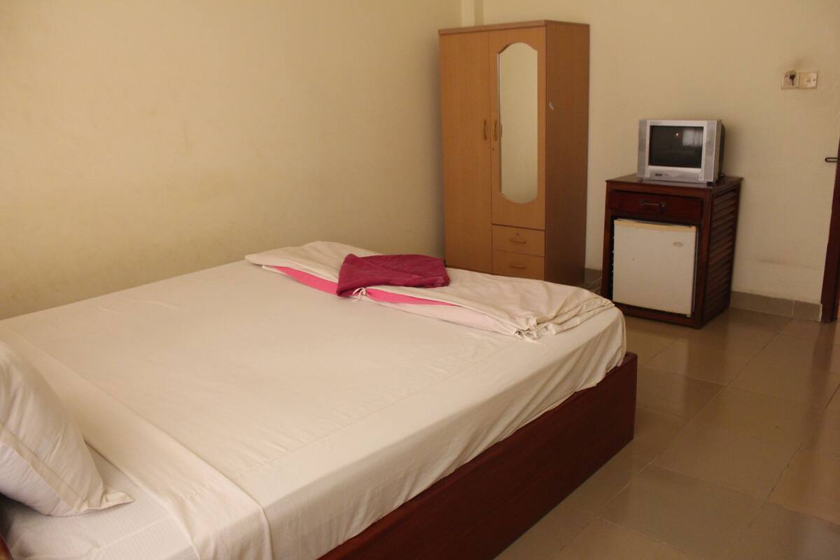 Una cama individual con colchón blanco y manta rosa, un armario de madera y un televisor pequeño sobre un mueble de madera.