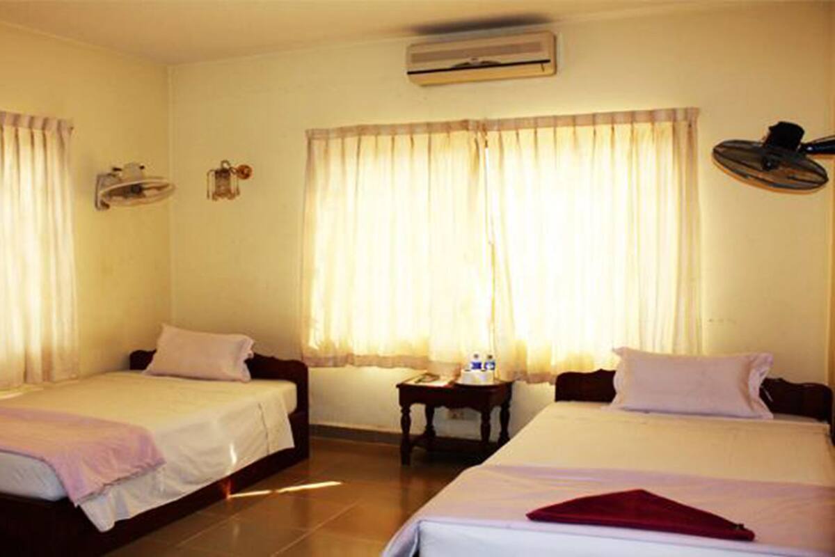 Habitación de hotel con dos camas individuales, una mesita pequeña, un aire acondicionado y una ventana con cortinas.