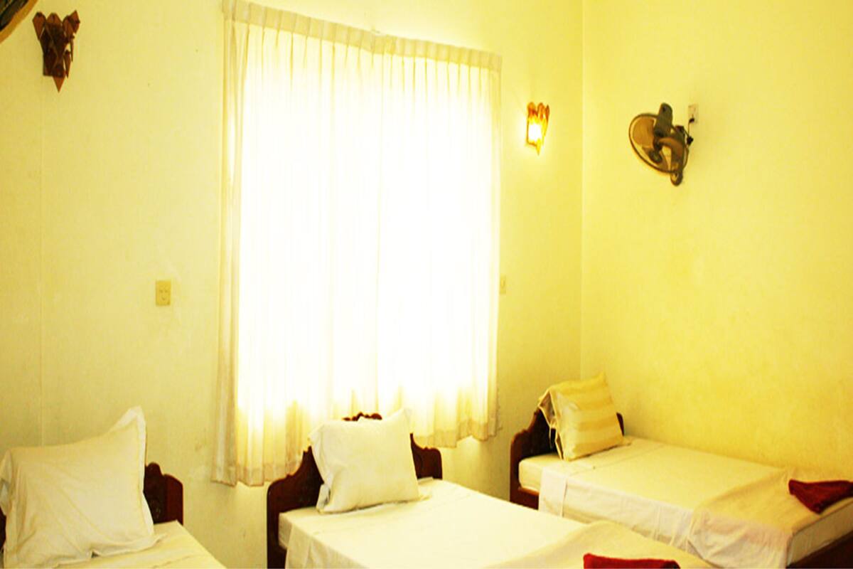 Una habitación con dos camas individuales, una ventana con cortinas translúcidas, una lámpara fijada a la pared y un pequeño adorno mural.