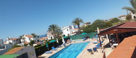 A swimming pool with lounge chairs and umbrellas, surrounded by buildings and palm trees.