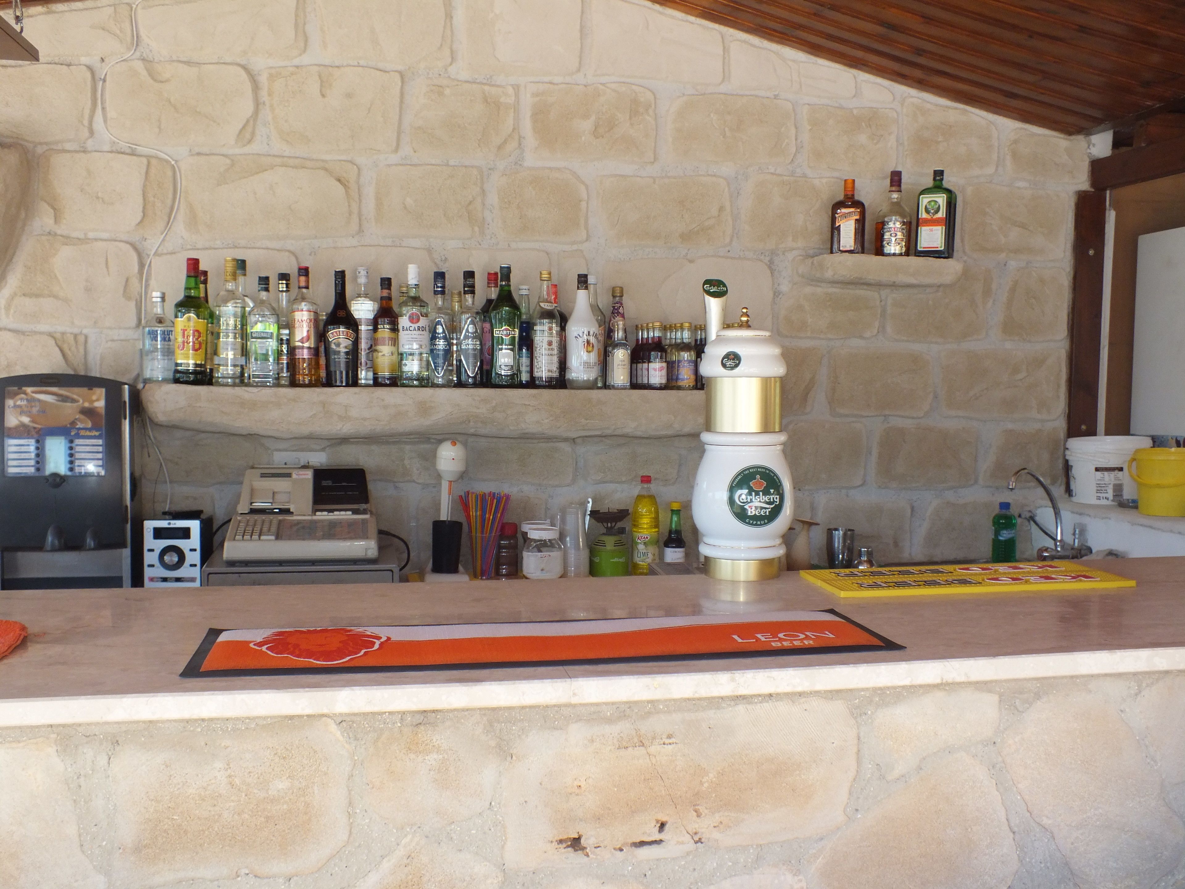 Een bar met een stenen muur, planken vol verschillende flessen, een biertap en een kassa.