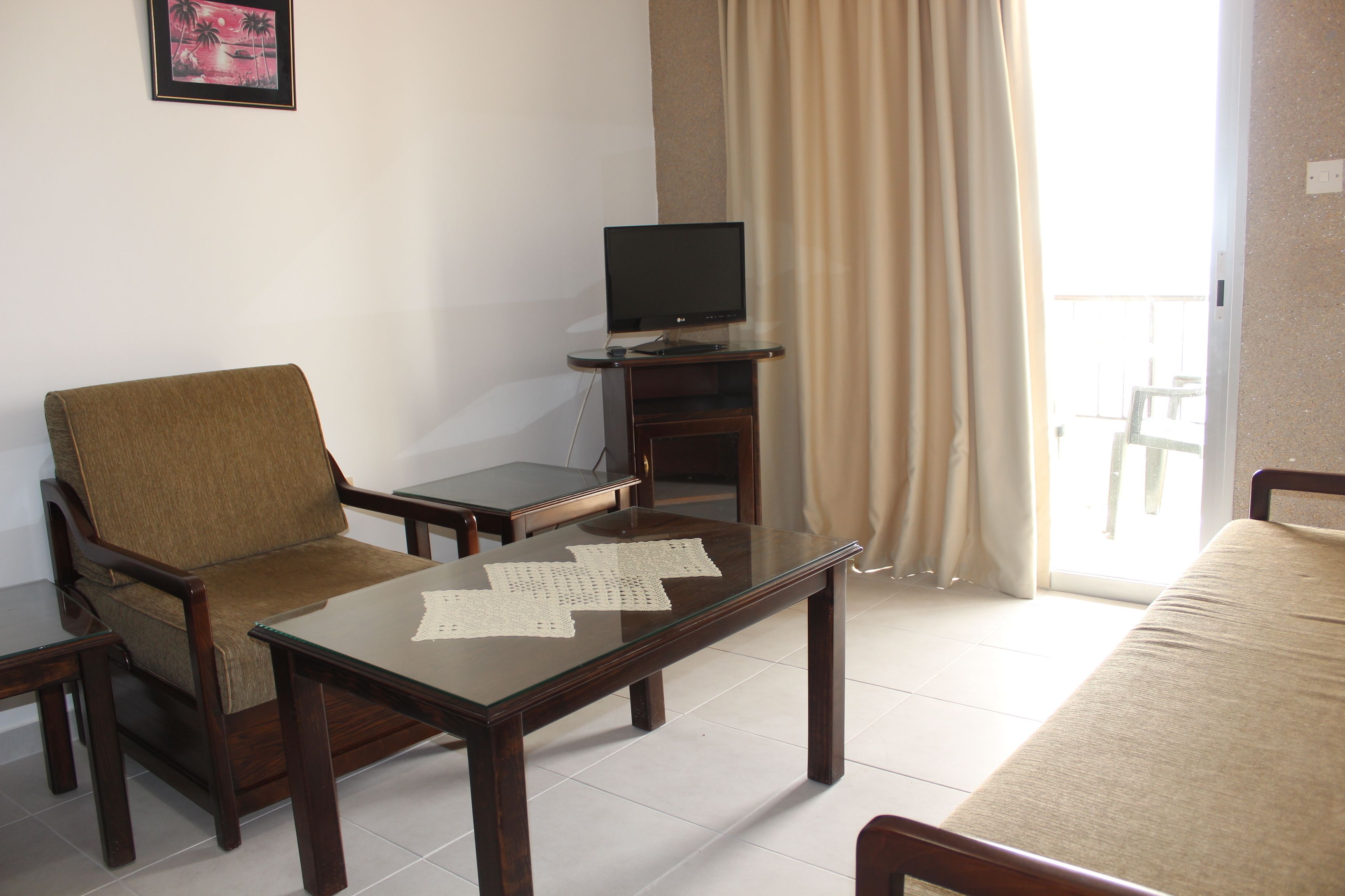 A hotel room with a brown armchair, a glass-top table with papers, a television on a stand, and a bed.