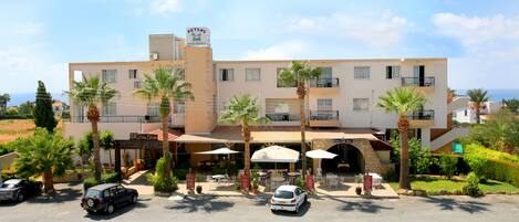 A hotel with a sign 'BETAS Apartments' and a 'Geralstar' restaurant. There are palm trees and a parking lot with cars.