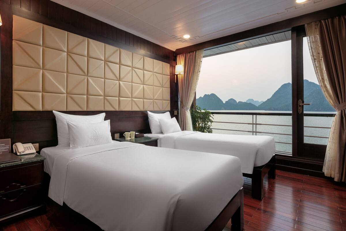 A hotel room with two beds, a wooden headboard, a nightstand with a phone, and a view of the water and mountains.