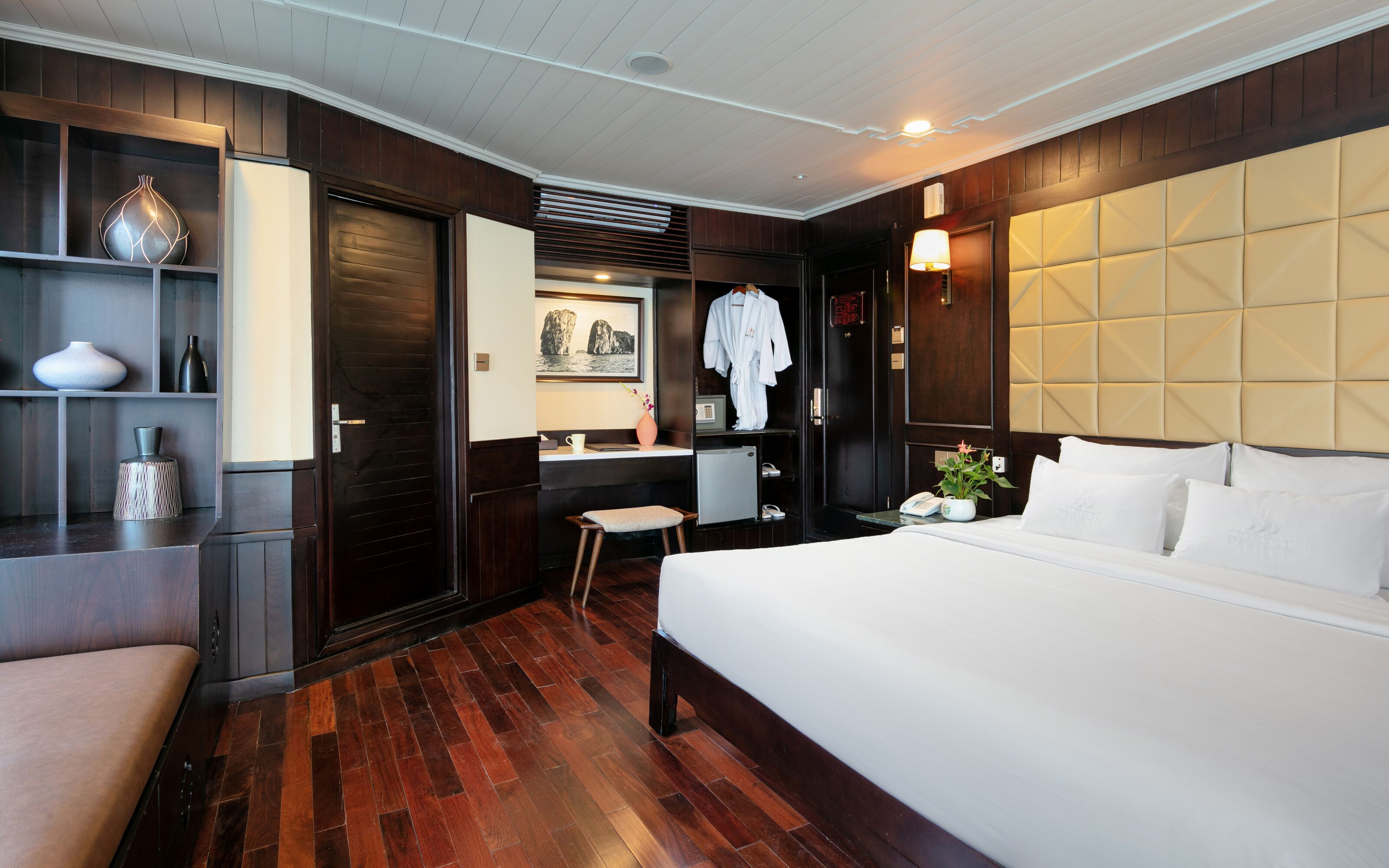 A hotel room with a large bed, a wooden floor, a vanity with a mirror, a small desk, and a shelf with decorative items.