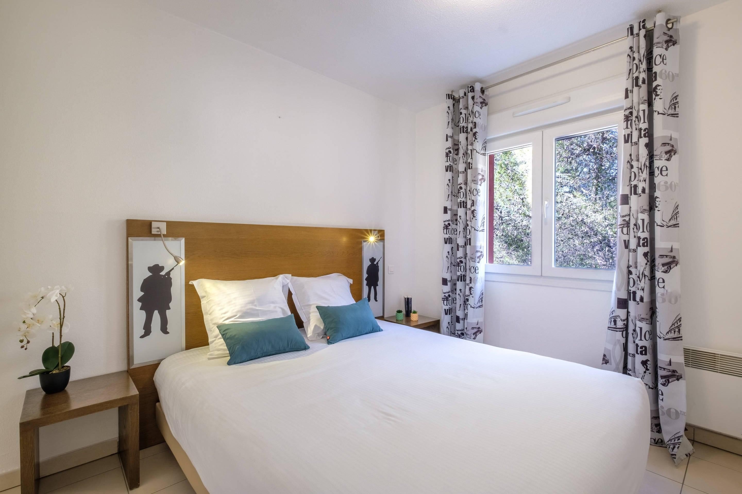 Une chambre à coucher avec une tête de lit en bois, un lit recouvert de draps blancs et agrémenté de deux oreillers, une table de chevet avec une plante, et une fenêtre ornée de rideaux à motifs.