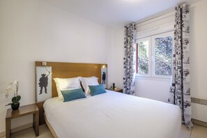 A bedroom with a wooden headboard, white bedding, a nightstand with a plant, and patterned curtains. - Comfort Aparthotel Antibes Le Maestria (Antibes)